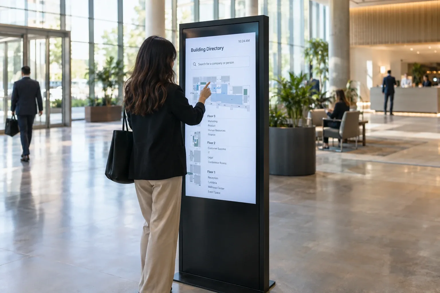 A visitor using a freestanding touch screen kiosk in an office lobby to search a building directory and floor map.