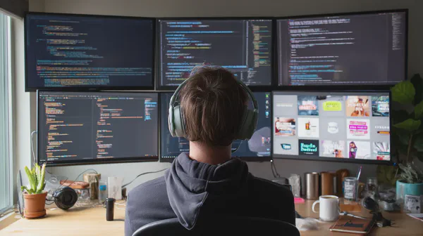 A developer, headphones on, sits before six bright monitors filled with code and vibrant designs, surrounded by plants and coffee mugs. It’s a focused, creative space for building custom digital signage apps.